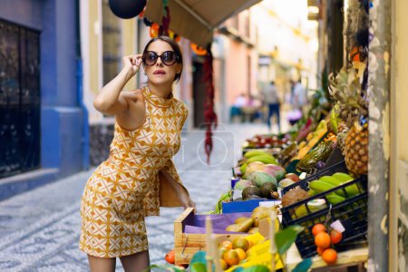 Photo for Young female model in stylish yellow dress standing and leaning towards stall with assorted fruits in street market while touching eyeglasses and looking at camera - Royalty Free Image
