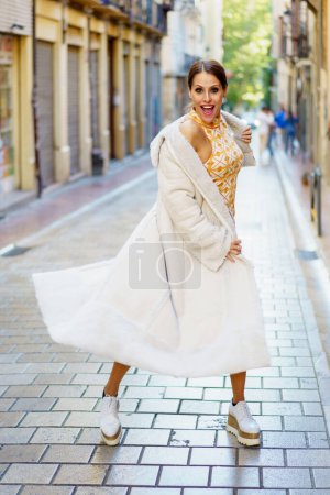 Foto de Sonriente mujer joven elegante con abrigo elegante y zapatos de pie en la calle pavimentada contra edificios viejos borrosos y mirando a la cámara - Imagen libre de derechos