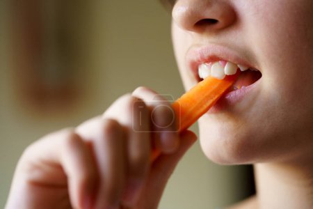 Photo for Closeup of crop unrecognizable teenage girl eating fresh healthy carrot slice at home - Royalty Free Image