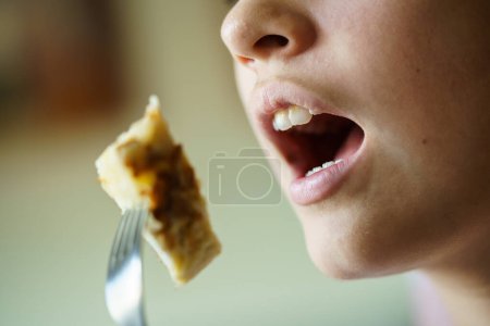 Foto de Primer plano de la cosecha irreconocible adolescente con la boca abierta a punto de comer tortilla de patatas deliciosa de tenedor en casa - Imagen libre de derechos