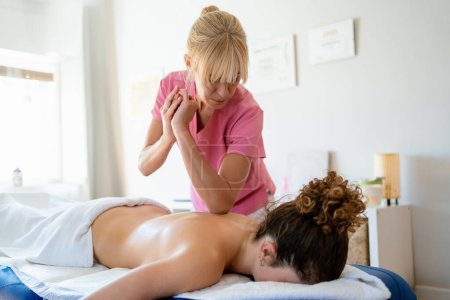Téléchargez les photos : Ostéopathe féminin concentré en uniforme en utilisant le coude tout en massant le dos du client seins nus couché sur le lit dans une clinique de physiothérapie moderne - en image libre de droit