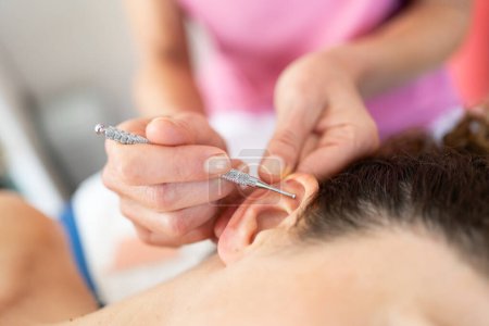 Téléchargez les photos : Crop cosmétique professionnel appliquant stylo de massage sur l'oreille de la femme pendant l'auriculothérapie dans le salon de beauté moderne sur fond flou - en image libre de droit