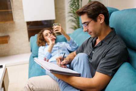 Photo for Side view of concentrated young man sitting on sofa and taking notes in notebook, while spending time with woman lying on couch with smartphone in living room - Royalty Free Image