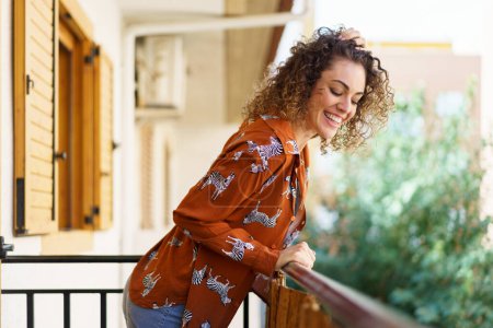 Photo for Side view of young curly haired happy female looking down while leaning on balcony railing at modern building against blurred background - Royalty Free Image