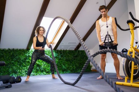 Photo for Full body of young sporty woman in activewear exercising with battle ropes during training near fitness buddy in contemporary gym with equipment - Royalty Free Image