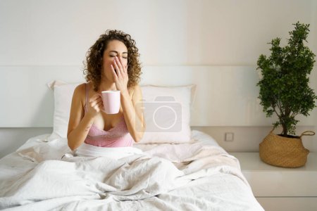 Photo for Young woman in nightwear yawning while holding coffee mug in bed during morning - Royalty Free Image