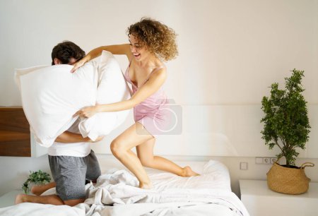 Photo for Cheerful young woman and man in sleepwear fighting with pillows on comfortable bed in bedroom at home - Royalty Free Image