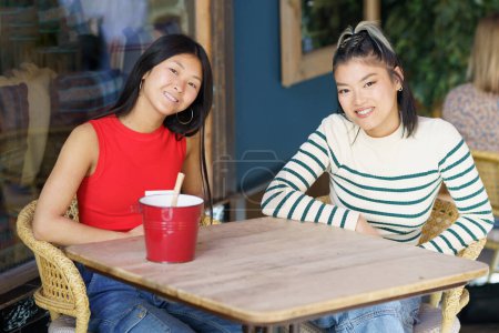 Téléchargez les photos : Souriant jeunes filles asiatiques en vêtements décontractés assis ensemble à la table en bois et discuter des plans tout en passant du temps dans le café - en image libre de droit