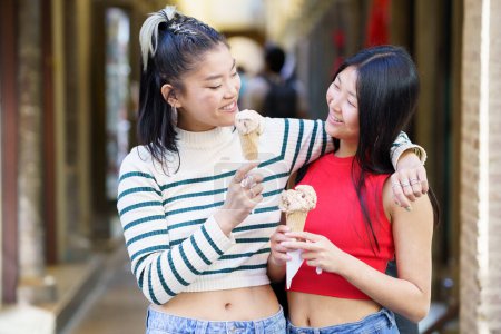 Photo for Cheerful Asian female standing with hand on shoulder of friend while eating delicious ice cream in street against blurred background - Royalty Free Image
