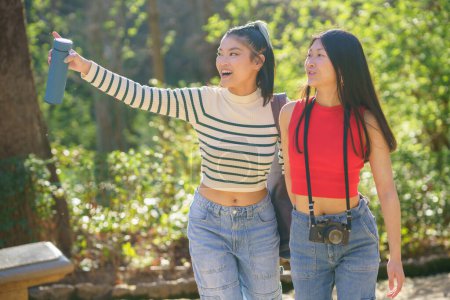 Photo for Cheerful young Asian female friends in casual apparel, standing in green forest of Alhambra and pointing away while enjoying picturesque views during summer trip in Granada - Royalty Free Image