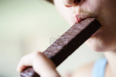 Foto de Primer plano de la cosecha anónima chica joven comiendo barra de proteína de chocolate en casa - Imagen libre de derechos
