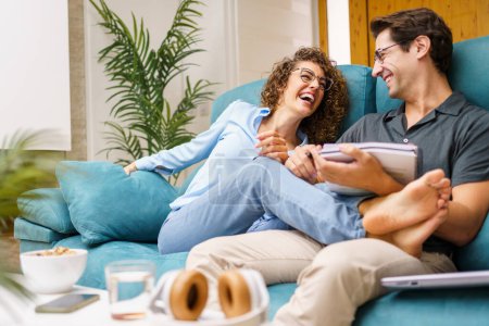 Photo for Happy young wife in casual clothes sitting with legs stretched on husband with notebook while laughing bonding together over sofa in cozy living room - Royalty Free Image