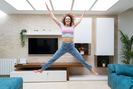 Téléchargez les photos : Jeune femme excitée aux cheveux bouclés en vêtements décontractés sautant au-dessus du sol et levant les bras tout en s'amusant à la maison - en image libre de droit
