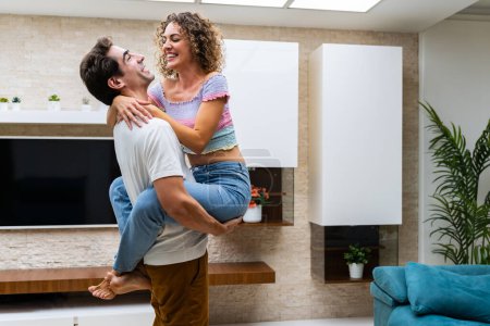 Photo for Side view of cheerful young couple hugging while standing against TV and shelves in modern apartment with potted plants on wall at home - Royalty Free Image