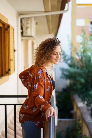Photo for Side view of young woman in casual clothes with curly hair standing on balcony and looking away - Royalty Free Image