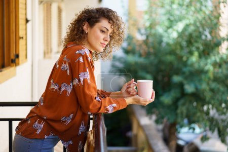 Photo for Side view of young curly haired female wearing casual clothes with coffee cup standing against blurred background in modern building - Royalty Free Image