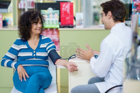 Foto de Farmacéutica competente que consulta a una paciente durante la medición de la presión arterial mientras está sentada en la farmacia y se mira entre sí - Imagen libre de derechos