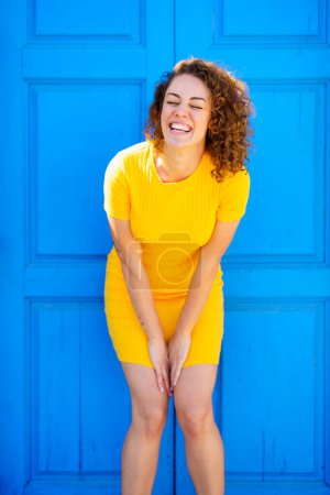Photo for Delighted young female in yellow dress with curly brown hair bending forward and smiling while laughing with closed eyes against blue building on street - Royalty Free Image