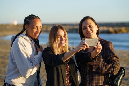 Téléchargez les photos : Copines multiethniques positives souriant joyeusement tout en prenant autoportrait sur téléphone portable sur la côte sablonneuse de la mer au coucher du soleil - en image libre de droit