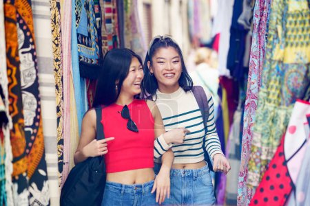 Foto de Sonriendo a las jóvenes amigas en ropa casual caminando por la colorida tienda de ropa en el mercado callejero de la ciudad durante las vacaciones de verano - Imagen libre de derechos