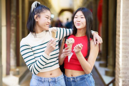 Téléchargez les photos : Femme asiatique positive debout avec la main sur l'épaule d'un ami tout en mangeant de délicieuses glaces dans la rue sur fond flou - en image libre de droit