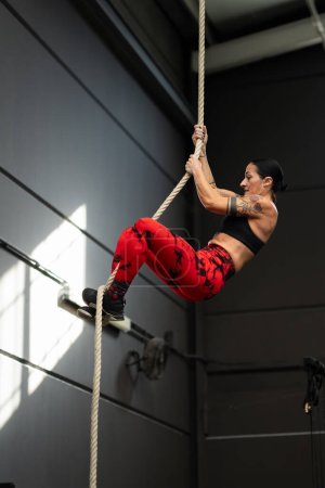 Photo for Vertical photo of a strong mature woman with tattooed arms climbing a rope in a cross training gym - Royalty Free Image