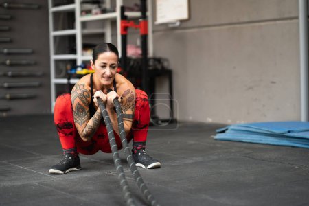 Foto de Mujer fuerte en cuclillas usando una cuerda de batalla para trabajar en un gimnasio - Imagen libre de derechos