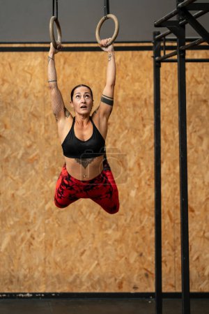 Vertikales Foto einer sportlichen Frau, die an olympischen Ringen hängt, während sie in einem Fitnessstudio trainiert