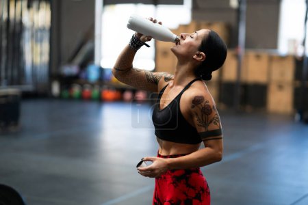 Téléchargez les photos : Vue latérale d'une femme en forme d'eau potable forment une bouteille réutilisable dans une salle de gym - en image libre de droit