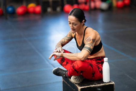 Téléchargez les photos : Sportswoman reposant dans la salle de gym en utilisant la montre intelligente assis sur une boîte - en image libre de droit