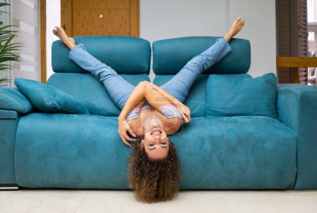 Téléchargez les photos : Joyeux jeune femme en jeans et haut avec les cheveux bouclés couché à l'envers sur le canapé dans le salon et s'amuser à la maison tout en appréciant - en image libre de droit