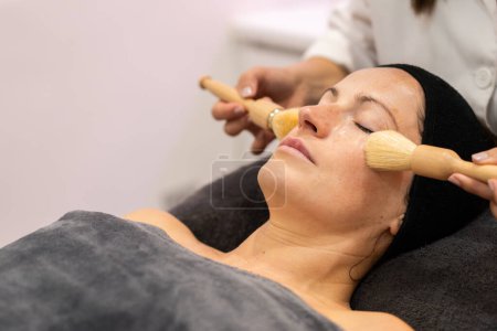 Photo for Crop beautician applying cosmetic cream on face of female customer with brushes during skin treatment in modern beauty center - Royalty Free Image