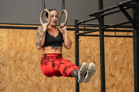 Téléchargez les photos : Femme effectuant des exercices de base suspendus à des anneaux olympiques dans un gymnase d'entraînement croisé - en image libre de droit