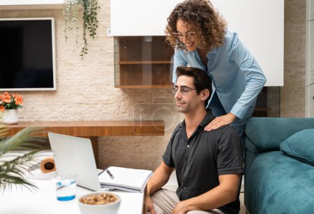 Téléchargez les photos : Joyeux jeune femme donnant massage des épaules à partenaire assis près de la table avec ordinateur portable et ordinateur portable dans le salon à la maison - en image libre de droit