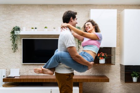 Photo for Side view of cheerful young couple hugging and looking at each other while smiling in modern living house with potted plants near tv on wall - Royalty Free Image