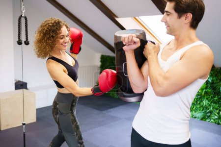 Foto de Joven luchadora sonriente en guantes de boxeo golpeando bolsa pesada durante el entrenamiento intenso con instructor personal en el centro deportivo - Imagen libre de derechos