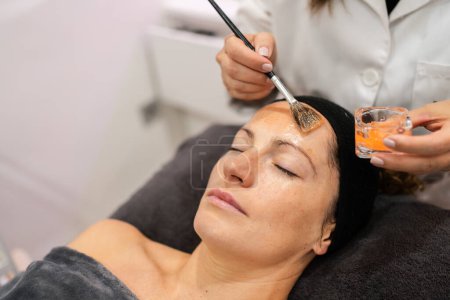 Photo for High angle of anonymous beautician with brush applying moisturizing treatment on face of relaxed woman from container in beauty salon - Royalty Free Image