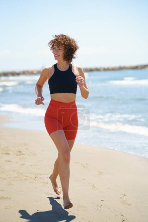 Téléchargez les photos : Corps complet de jeune femme souriante pieds nus en vêtements de sport, avec les cheveux bouclés jogging sur la plage de sable et regarder loin tout en se réchauffant près de la mer ondulante dans la journée ensoleillée d'été - en image libre de droit
