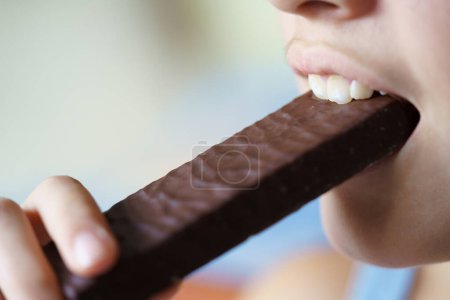 Foto de Primer plano de la cosecha anónima adolescente comiendo deliciosa barra de proteína de chocolate en casa - Imagen libre de derechos