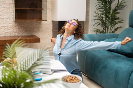 Téléchargez les photos : Jeune femme pigiste dans les lunettes et smart casual étirement des bras tout en travaillant sur ordinateur portable à la table dans le salon à la maison - en image libre de droit