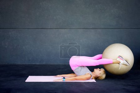 Téléchargez les photos : Vue latérale d'une jeune femme en forme flexible allongée sur un tapis et faisant des exercices de pilates avec une balle de fitness pendant l'entraînement au gymnase contre un mur gris pendant l'entraînement - en image libre de droit