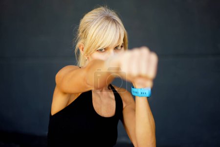 Téléchargez les photos : Combattant féminin aux cheveux blonds forts en débardeur noir dans un gymnase sombre et pratiquant des coups de poing pendant l'art martial, entraînement sur fond gris en regardant la caméra - en image libre de droit