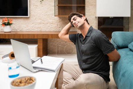 Photo for Happy young male freelancer in casual clothes stretching neck while sitting near laptop and notebook in living room at home - Royalty Free Image