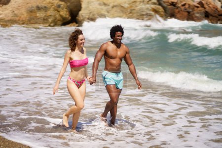 Photo for Full body of cheerful multiracial couple holding hands and strolling together on coast while enjoying summer trip in sea - Royalty Free Image