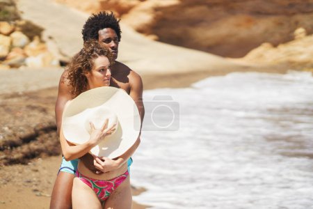 Photo for Young African American man with Afro hairstyle hugging girlfriend from behind standing on sandy beach, covering breast with hat while spending time together near waving sea - Royalty Free Image