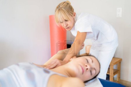 Téléchargez les photos : Physiothérapeute féminine ciblée en uniforme blanc massant les aisselles de la femme allongée sur le canapé pendant la séance d'ostéopathie - en image libre de droit