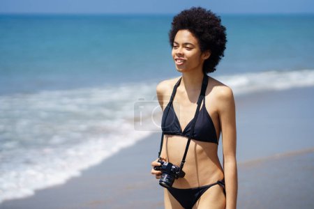 Foto de Mujer afroamericana optimista en traje de baño con cámara fotográfica profesional de pie en la orilla del mar mojada cerca del mar ondulante en un resort tropical con clima soleado - Imagen libre de derechos