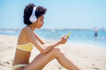 Téléchargez les photos : Vue latérale du contenu jeune ajustement afro-américaine millénaire avec les cheveux bouclés, dans les maillots de bain à la mode navigation smartphone, et écouter de la musique dans les écouteurs assis sur la plage de sable près de la mer - en image libre de droit