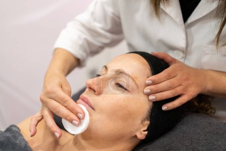 Téléchargez les photos : Crop cosmétique nettoyage visage de jeune cliente avec coton pendant le traitement de la peau dans le salon de beauté moderne - en image libre de droit
