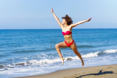 Photo for Smiling carefree young slim female redhead in pink bikini raising arms and jumping with legs in air while looking away, in daylight against blurred sea and blue sky - Royalty Free Image